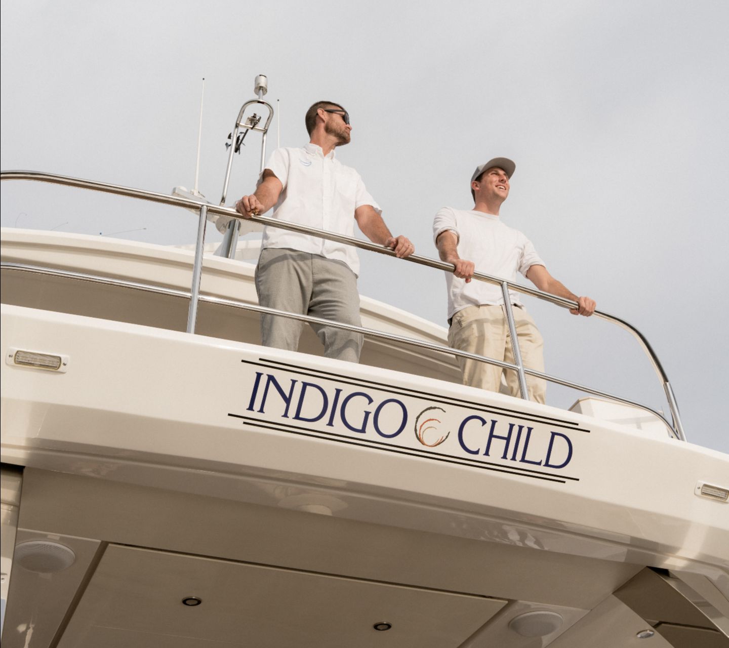 Two people stand on a yacht named Indigo Child under a cloudy sky.