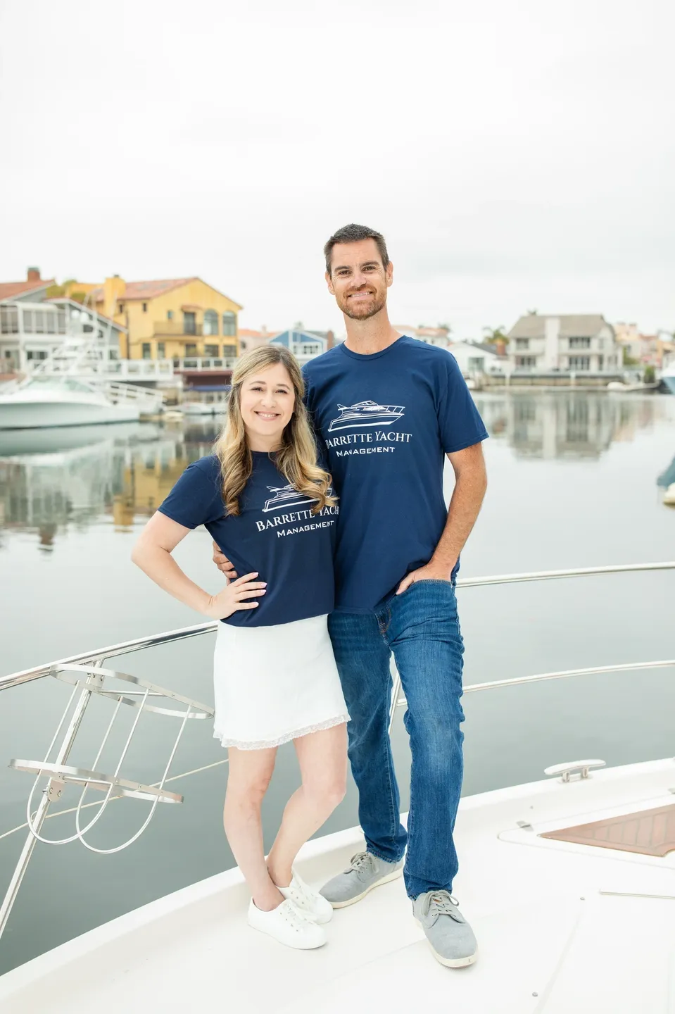 A smiling couple wearing matching navy blue shirts on a dock with boats and waterfront houses behind them.